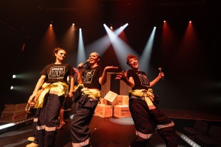 Dangerous Goods. Three black clad performers wear T shirts that say 'show up' and sing into microphones while spotlights shine down on them.