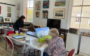 Opportunities. Grenfell Art Group, recipient of the latest CASP funding. Photo: Supplied. A painting studio with a table in the middle and artwork hang on the walls. A person can be seen on one side of the table, working on a landscape painting.