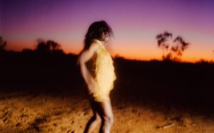 Still from the film ‘Marungka Tjalatjunu (Dipped In Black)’ by Derik Lynch and Matthew Thorne, showing as part of Queer PHOTO. Image: Courtesy of the artists. A figure wearing a gold dress with long hair, standing against a purple and orange sunset in a desert landscape.