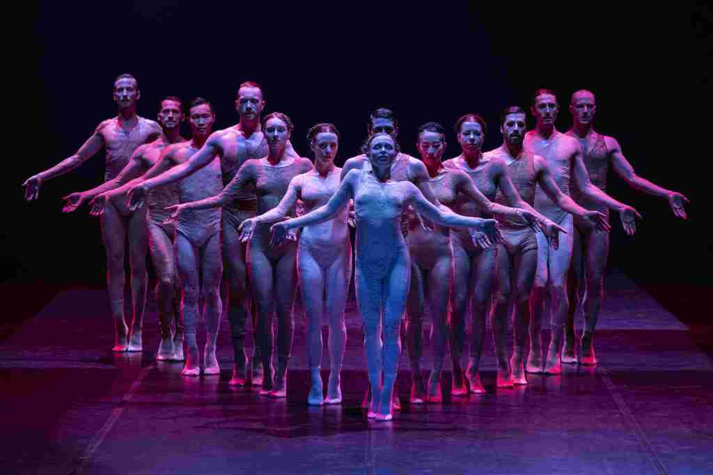 ‘SAABA’ by Sharon Eyal for GöteborgsOperans Danskompani. Image: Supplied. 13 dancers standing in a ‘V’ formation on a stage with purple and blue lighting, with their arms held out and on the tip of their toes. 