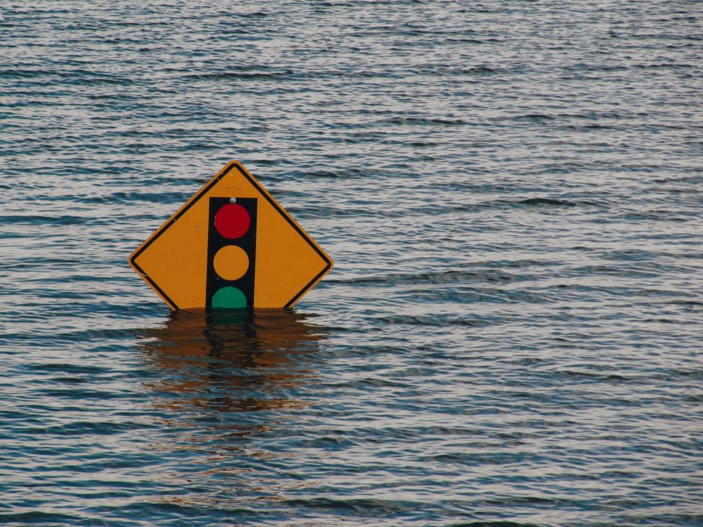 Photograph of a road sign warning of traffic lights ahead that is half-submerged in floodwaters.