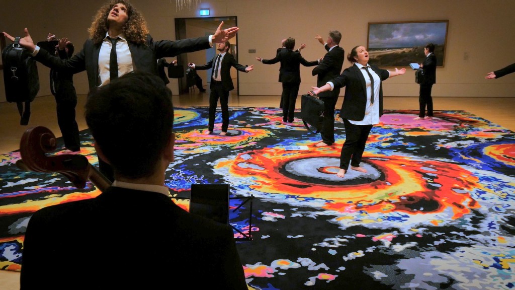 It Lights the Whole Sky. Image is a choral group spread through a gallery space, standing on a multicoloured rug surrounded by 19th century paintings of weather phenomena and storms, singing. They all wear black suits and ties, and white shirts.