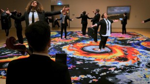 It Lights the Whole Sky. Image is a choral group spread through a gallery space, standing on a multicoloured rug surrounded by 19th century paintings of weather phenomena and storms, singing. They all wear black suits and ties, and white shirts.