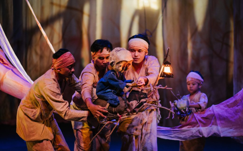 ‘A Bucket of Beetles’ at Sydney Festival 2024. Photo: Rendha Rais & Rangga Yudhistira. Performers dressed in earth-coloured clothing holding a small puppet in the middle.