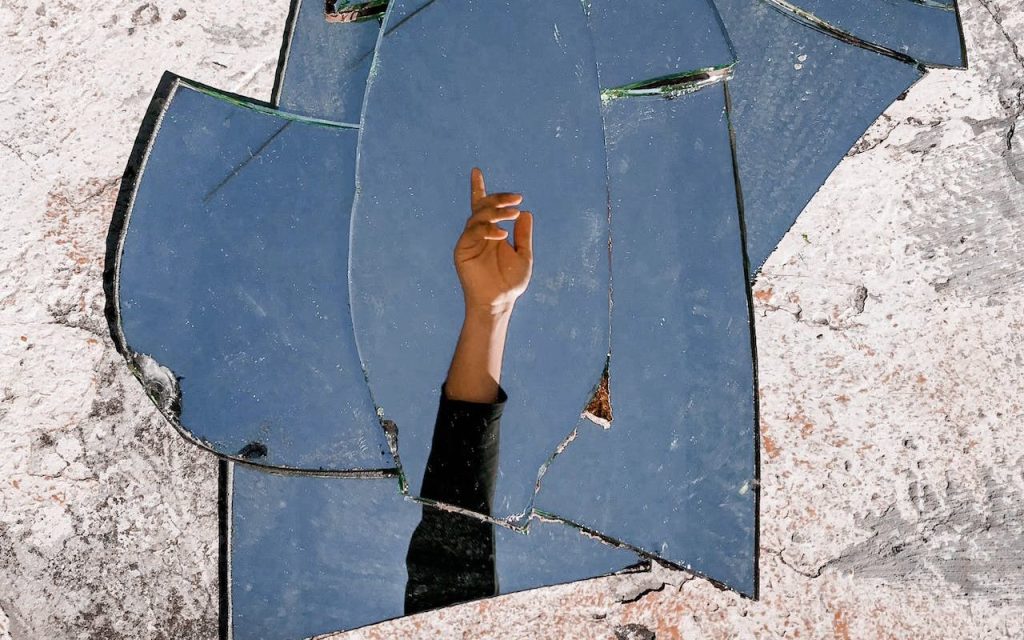 Photo: Bruno Pires on Pexels. Photo of a mirror shattered on a gray surface with the reflection of a person's hand.