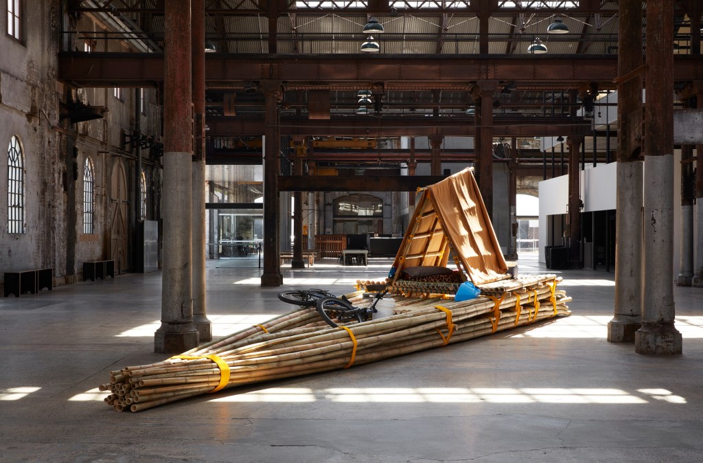 Salote Tawale, ‘No Location’, 2021. Installation view, ‘I remember you’, Carriageworks, 2023. Purchased 2021 with funds from the Bequest of Jennifer Taylor through the Queensland Art Gallery l Gallery of Modern Art Foundation. Collection: QAGOMA. Photo: Zan Wimberley. A huge raft mad of bamboo sits in a grey industrial space with sunlight. 
