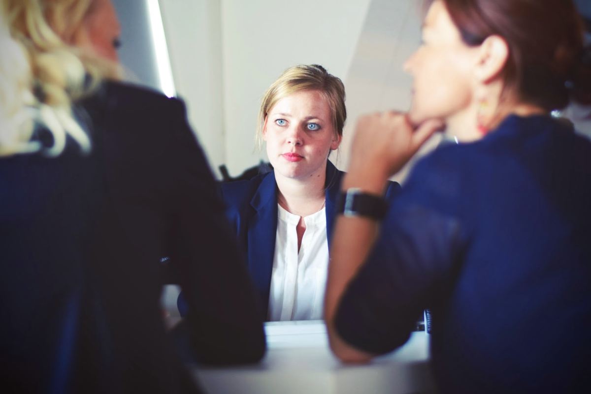 job interview. Three women at a desk, two with their backs to the camera, one on the other side being interviewed.