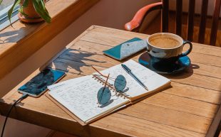 Photo: Toa Heftiba on Unsplash. Sun shines through the window of a streetside cafe. A cup of coffee, phone, sunglasses and a notebook is spread open on a wooden table.