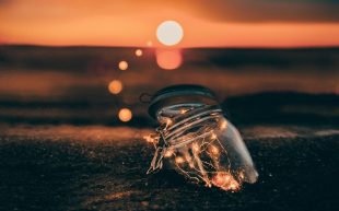 Photo: Yeshi Kangrang on Unsplash. A glass jar filled with fairy lights half burried into the sand at the beach against a setting sun.