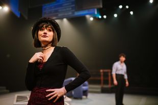 Billy Fogarty, a non-binary actor, performing on stage. Image is a performer downstage in a black top and black hat, with one hand on their hip and the other touching their lower neck. Behind them in the background is a figure in dark trousers and a light shirt.