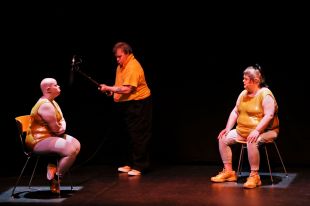 Two women sit in chairs on a bare stage facing one another. A man stands between them with a boom microphone.