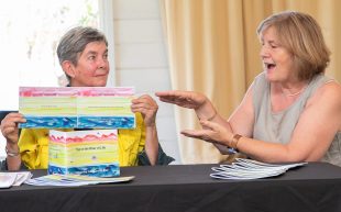 L-R: Project Alchemy artist Sue Norman and Colleen Weir, collaborators on 'Nye River of Life'. Norman has short grey hair and she is holding up the cover of her book, featuring rows of different colours. Weir is speaking and gesturing towards the book. They are booth sitting down behind a table covered with black cloth.