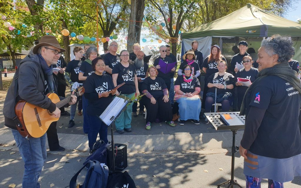 With One Voice choir members at the National Folk Festival 2023. Photo: With One Voice Australia. A group of choir members with diverse skin tones sitting and standing outside in front of a festival scenery with trees. A person is playing a guitar and another is conducting.