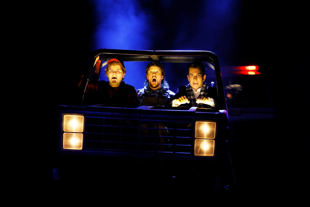 Groundhog Day the Musical. Three men on stage in the dark sitting in a car looking out the windscreen at the audience.