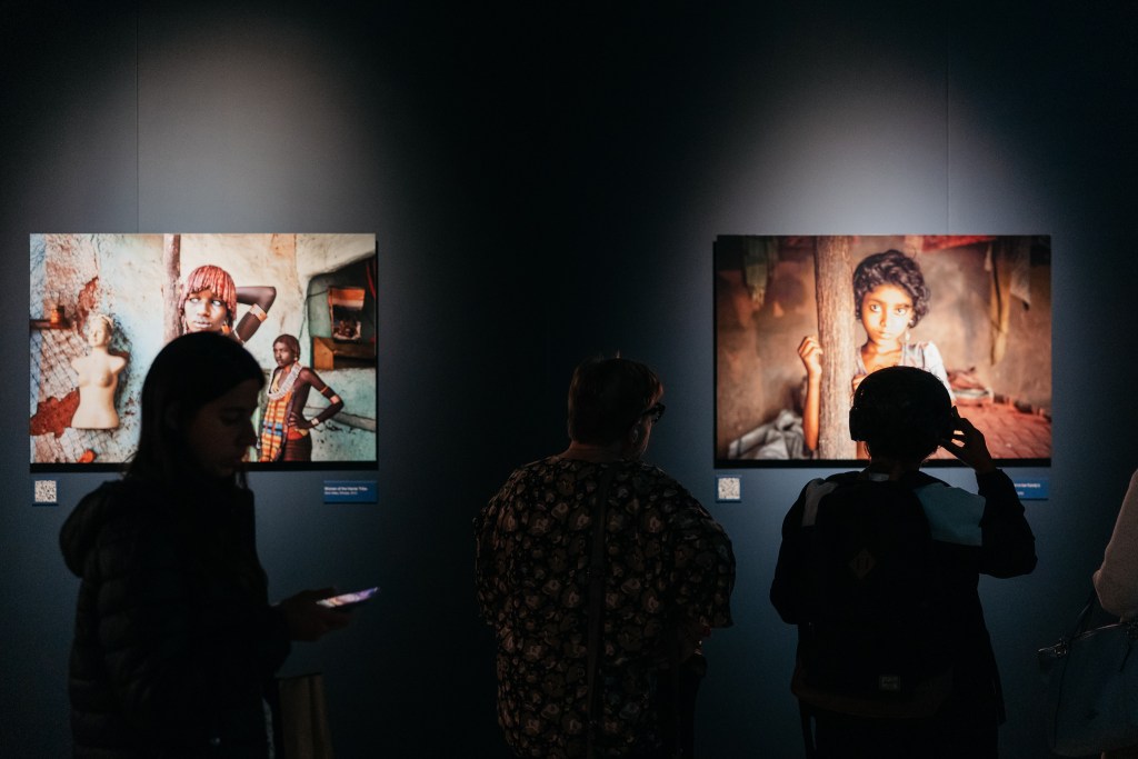 Two exhibition visitors regard two well-known photographs by Steve McCurry in a dark gallery space.