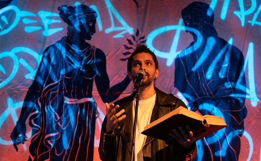 Greek-Australian poet and spoken word performer Luka Lesson gestures with his right hand while holding a book in his left.