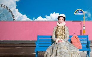 A woman in early 19th century garb sits at a Brisbane bus stop. The Wheel of Brisbane Ferris Wheel is visible behind her.