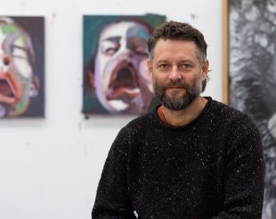 Man with beard and black jumper and paintings in background. Ben Quilty.