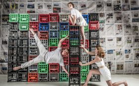 Three young circus performers wearing clean white clothing perform acts on a stack of milk crates. One is holding the crates while another is performing the human flag, and a third is climbing on top of the tower. In the background is a wall of crates in black, green, red and blue, with the back wall covered with newspapers.
