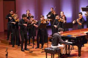 A man playing the piano is on the right hand side, in the background surrounding him are violinists, standing upright. Camerata, Alex Raineri.