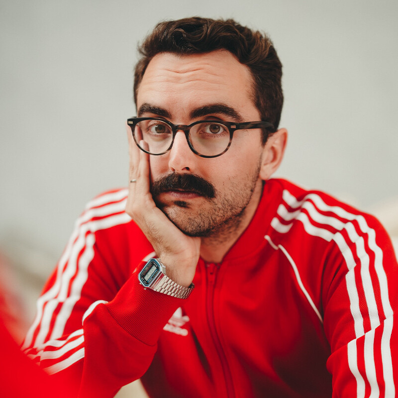 Blinded by the Whites. Image is a man in glasses with a thick moustache wearing a red and white striped track suit top and resting his chin in his palm.