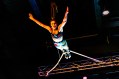 A young female circus performer flies through the air, suspended by her feet from a rope. She has a long plait which accentuates her movement as it too swings through the air.