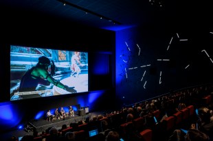 Future of Arts, Culture & Technology Symposium 2024 at ACMI, ‘Presenting the new canon in 2050’ panel. Photo: Gianna Rizzo. People sitting in a dark cinema with four people sitting on stage below a large screen showing a digital work (still). The work features a human figure moving their upper body with arms outstretched.