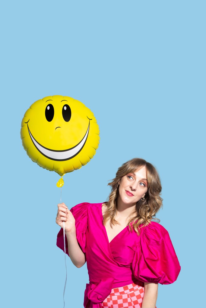 Gillian Cosgriff. Image is a blue background and a woman holding a yellow smiley face balloon and wearing a pink blouse. She has shoulder length fair hair.