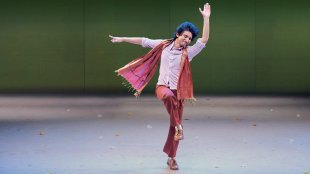 A curly-headed young man in traditional Sri Lankan dress dances joyfully on a bare stage.