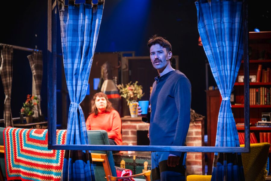 Logue Lake: a male actor holding a coffee mug, standing on stage framed by an open window with curtains at the side.