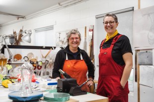 mentor. Two women stand side by side in an artist's studio. They are looking at the camera and smiling, wearing black tops with orange/red aprons over the top.