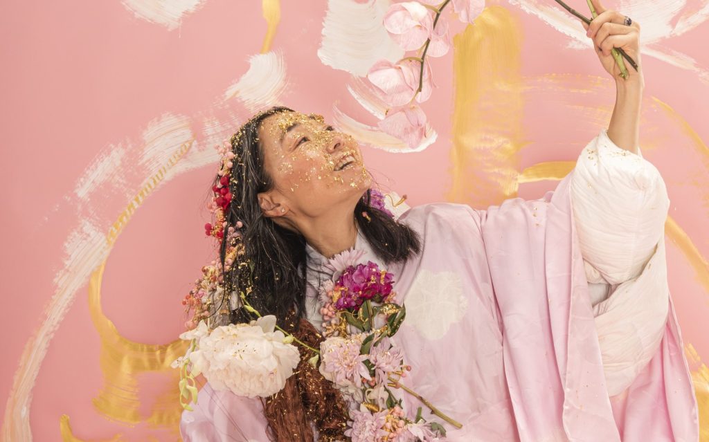 An Asian woman in her late 40s covered in dry flowers and gold glitter. She is holding an orchid branch with pink blossoms and smiling. The background appears to be an artwork with pink, white and gold abstract strokes.