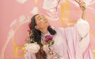 An Asian woman in her late 40s covered in dry flowers and gold glitter. She is holding an orchid branch with pink blossoms and smiling. The background appears to be an artwork with pink, white and gold abstract strokes.