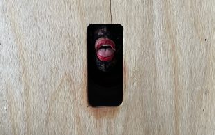 Emil Cañita. A gloryhole made from a timber board reveals a seductive half-opened mouth with red lipstick and framed by black lace.
