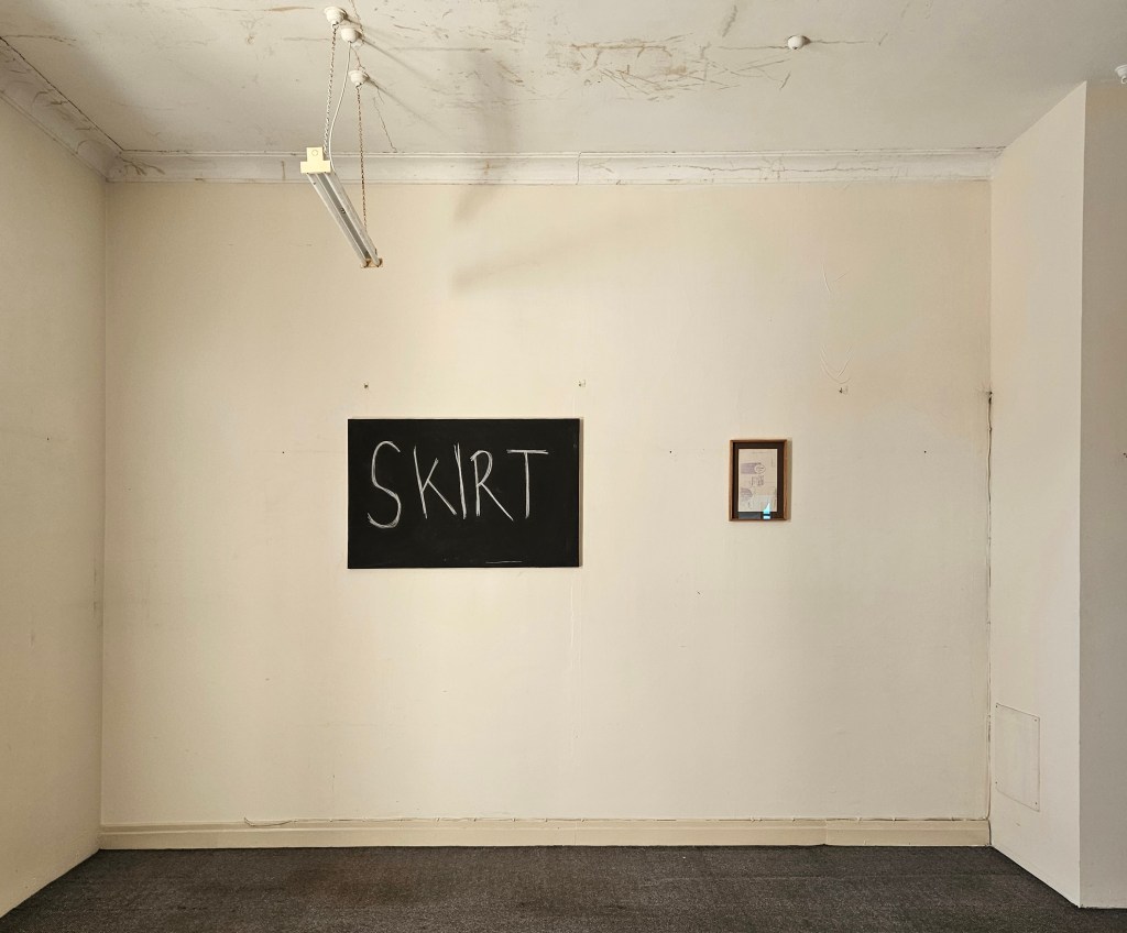 ‘Archie Moore: One Work From the Collection of Rebecca Ross and One From the Collection of Dirk Yates’, installation view at 1WORKROOM9. Photo: Supplied. Two small artworks hang on a yellow-ish wall, the one of the right has the word ‘SKIRT’ written in chalk on a blackboard. The work on the right is smaller and framed behind reflective surface, but illegible from this distance. The space feels rundown, with grey carpet and a ceiling with marks. 
