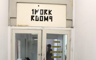 1WORKROOM9 occupies the previous space of Chinese Fraternity Association of Queensland. Photo: ArtsHub. A glass door with white timbre borders opens into a corridor. On the top of the door is a sign with ‘1WORKROOM9’, in the door itself is a row of letters stuck on, displaying ‘CHINESE FRATERNITY ASSOCIATION OF QUEENSLAND INC’