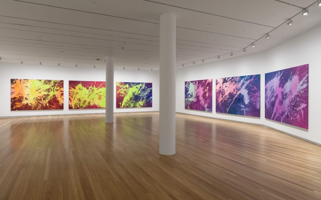Installation view ‘Ash Keating: PRESSURE’ at Bunjil Place. Photo: Michael Pham. A vast gallery space with polished timber floor and two white columns running through it. There are six large canvases with splashes of coloured paint, ranging from yellow, orange, red to green, blue and purple.