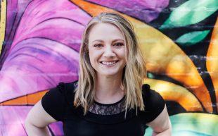 Emily Sheehan. Photo: Supplied. A young white woman with shoulder-length blonde hair and smiling at the camera. She is wearing a black tshirt and standing in front of a vibrant wall mural.