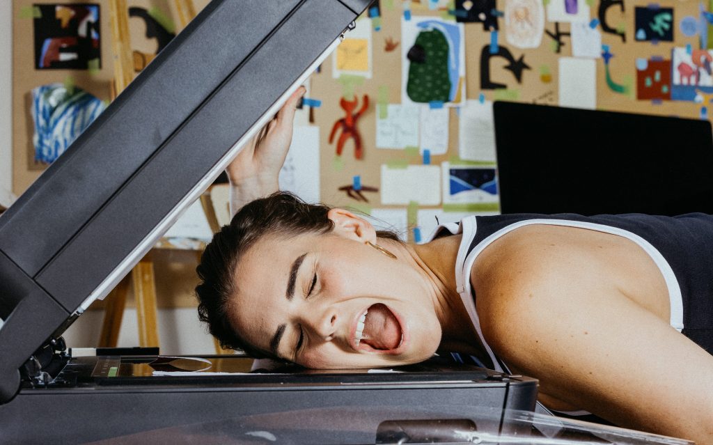 Eloise Lark, one of the artists participating in TasmanAi. Photo: Supplied. A playful photo of a women sticking her head on the scanning machine and making an exaggerated facial expression. Snippets of posters and artworks can be seen on a board behind her.