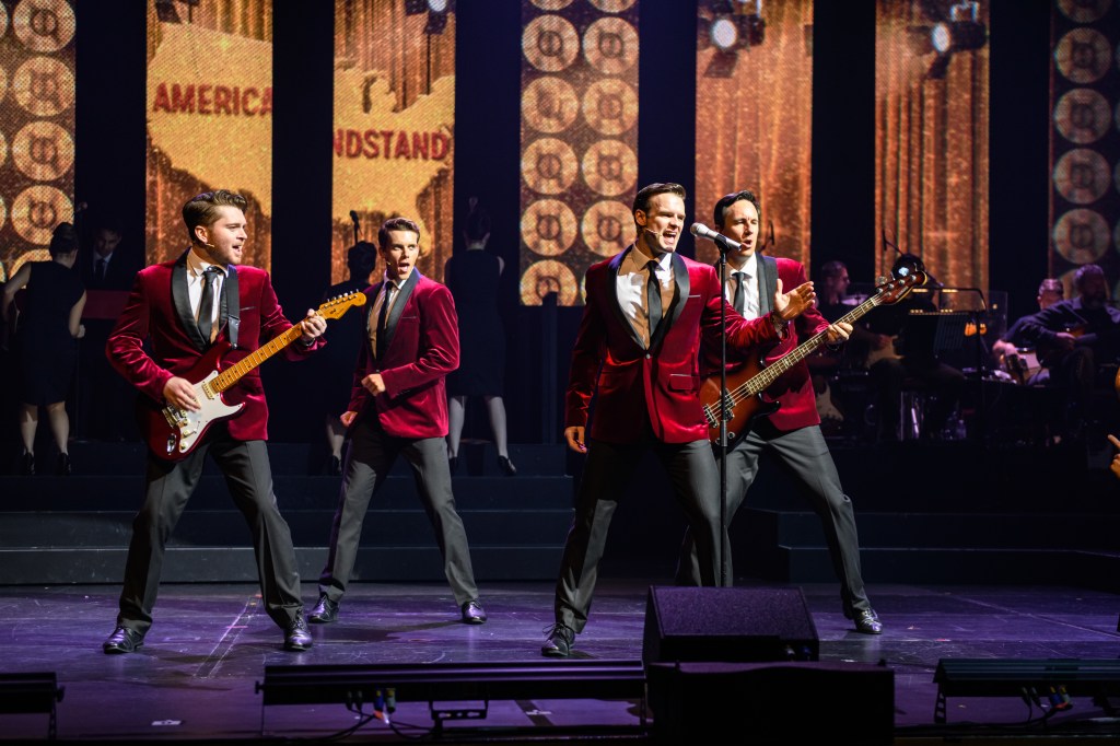 Four performers on stage wearing red velvet suits, white shirts, ties and black dress paints. Two of them are playing the guitar while the other two are singing. There are columns of screens in the background, projecting glittery gold imagery and the American map. 