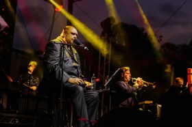 Mona Foma, Kutcha Edwards and the Australian Art Orchestra. Image is of a First Nations man singing at night on a stage, with a band of musicians around him and yellow spotlights beaming across the image.