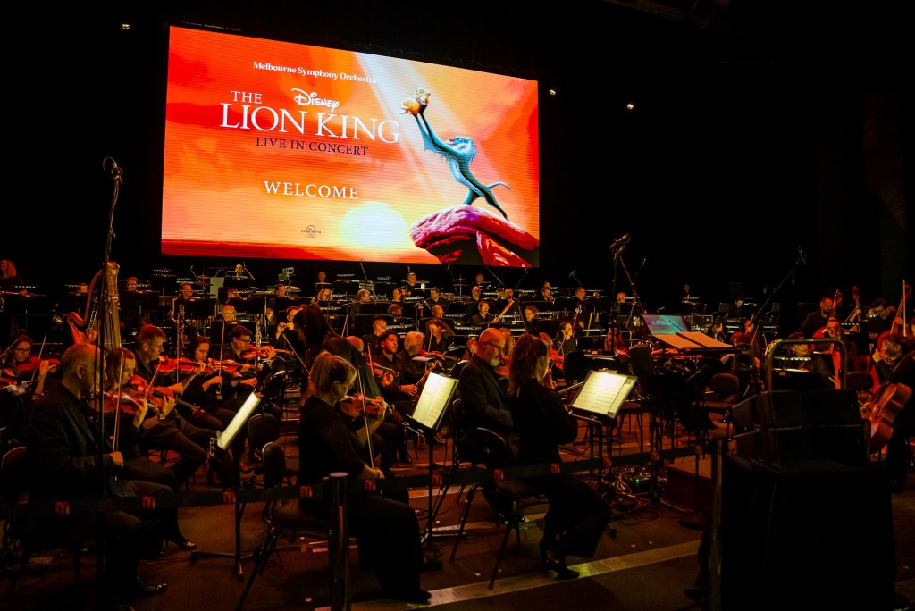 A screen displays the words The Lion King, in fornt of the screen is an orchestra
