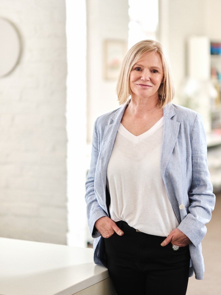 Blonde haired woman with blue jacket on standing with hands in pockets. Designer.