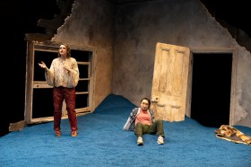 Meet Me at Dawn. Two women on a small stage set of a two sides of a broken down wall (with a door in one and window in the other), blue sand beneath. One woman is seated leaning back and the other is standing and gesturing with her palms face up.