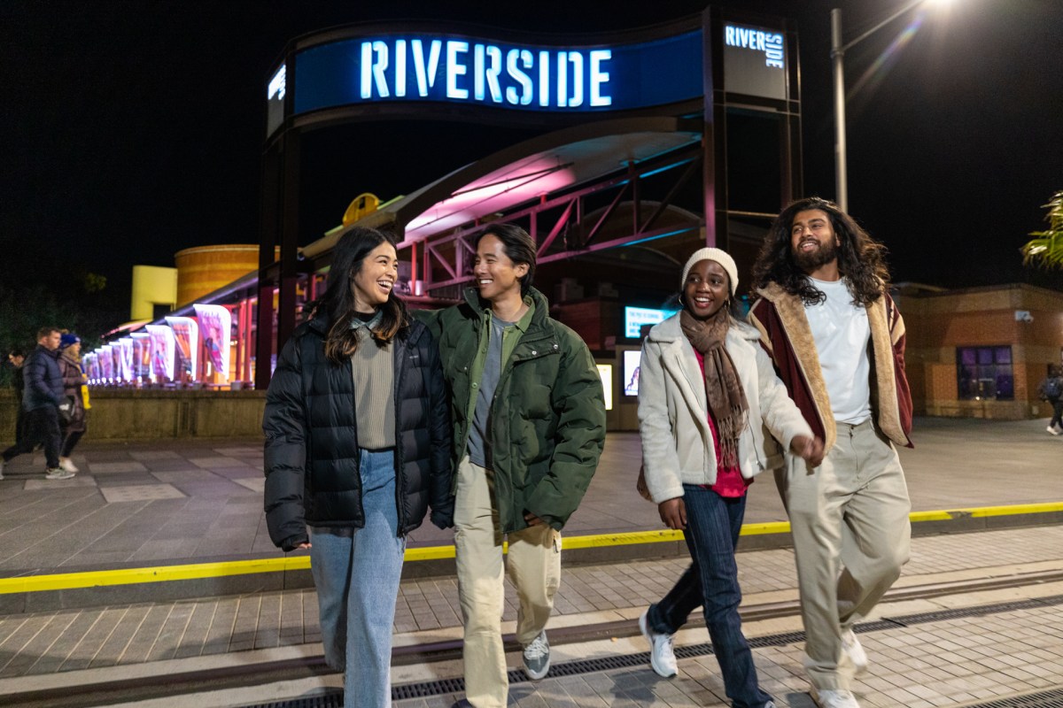 Riverside Theatres. Image is a group of four young people standing on the steps of a building that has the word RIVERSIDE in a blue neon sign at the top. They are looking at each other.