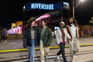 Riverside Theatres. Image is a group of four young people standing on the steps of a building that has the word RIVERSIDE in a blue neon sign at the top. They are looking at each other.