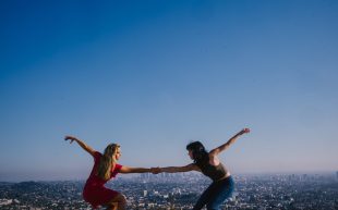 Benjamin Millepied's 'Romeo & Juliet' with female-female, male-male, and female-male couples. Photo: Josh Rose. Two female performers balance with holding hands with each other. They appear to be on a tall platform outdoors, overlooking a city scape and blue sky.