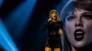 Taylor Swift: A picture of Taylor Swift in a little black dress holding a microphone, performing on stage in front of a projected image of her face.