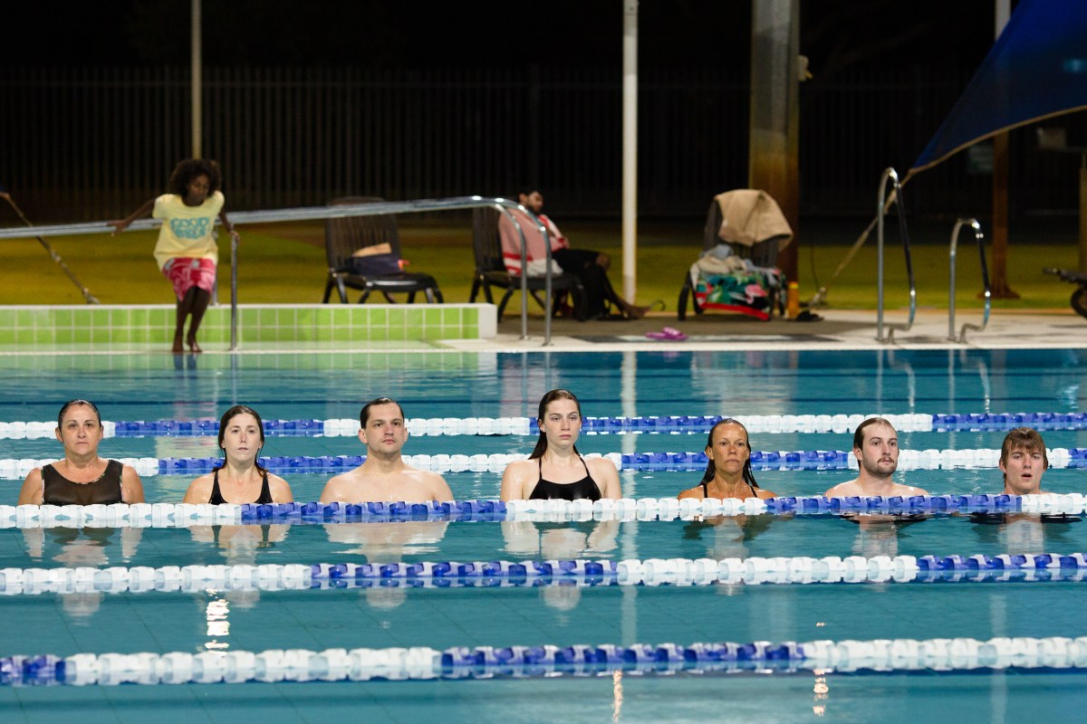 Seven figures are submerged in a pool. They are all lined up in one lane looking towards the side of the pool.