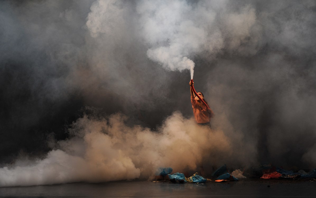Are we not drawn onward to a new erA. The image is of a single person wearing a nose/mouth mask on a stage holding up a smoking flare, surrounded by smoke with bits of detritus - fabric - at their feet.
